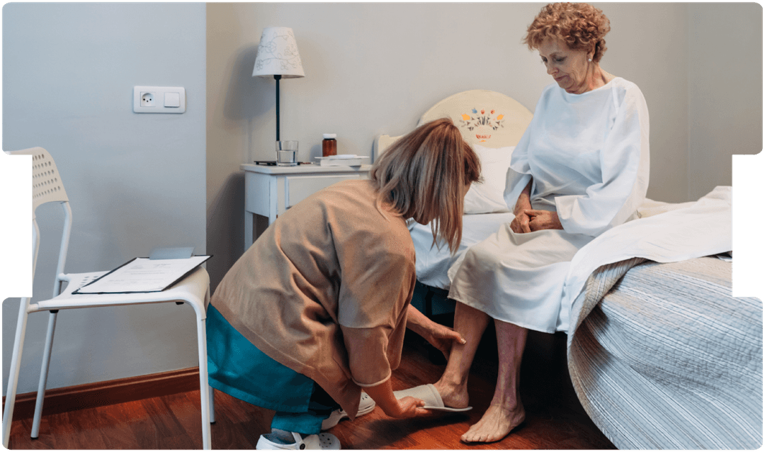 A caregiver gently helps an elderly woman with footwear beside her bed, reflecting compassionate elderly care, home care support, and patient dignity.