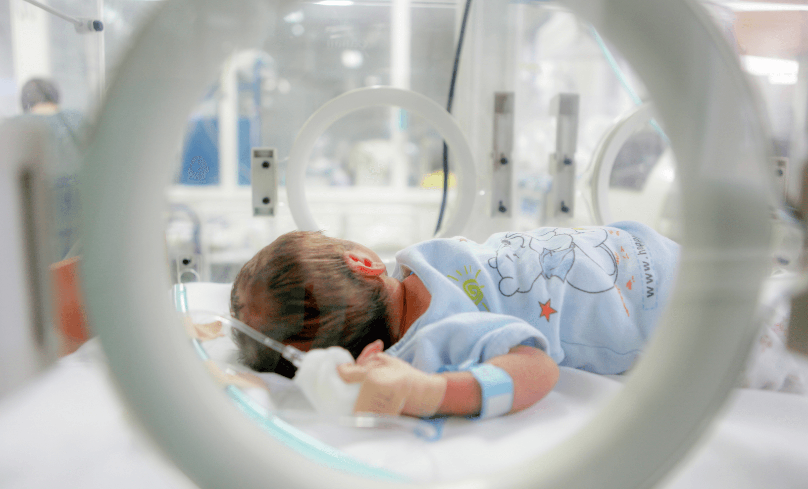 Newborn baby lying in a hospital incubator, wearing a blue onesie and monitored with medical tubes and wristband, highlighting neonatal intensive care in a clinical setting.