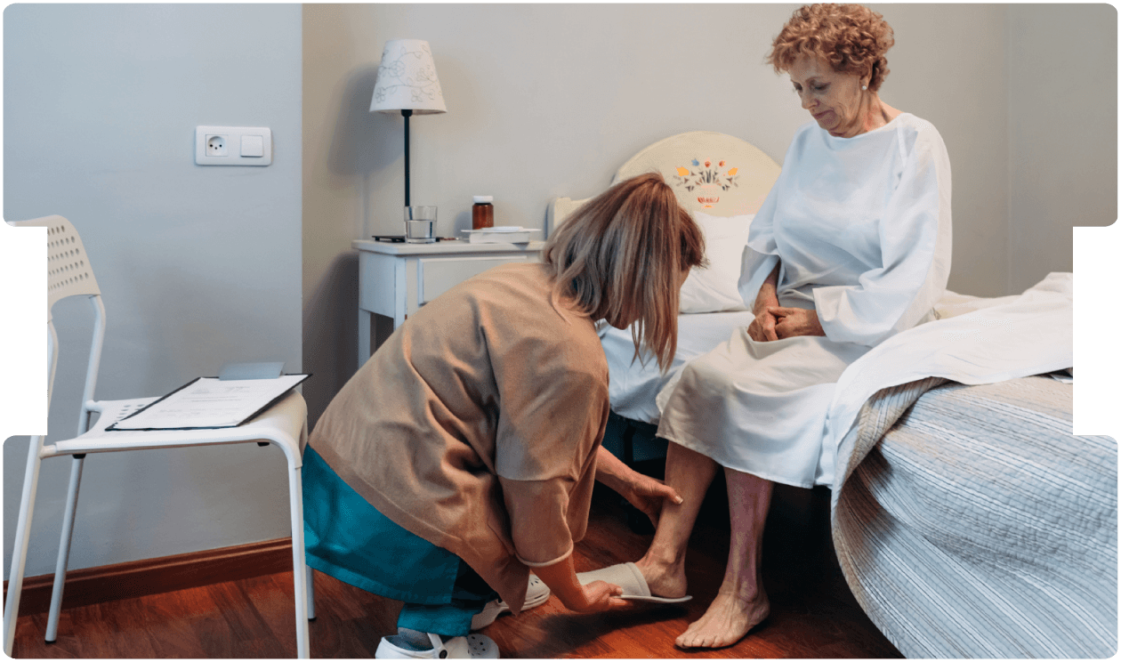A caregiver gently helps an elderly woman with footwear beside her bed, reflecting compassionate elderly care, home care support, and patient dignity.